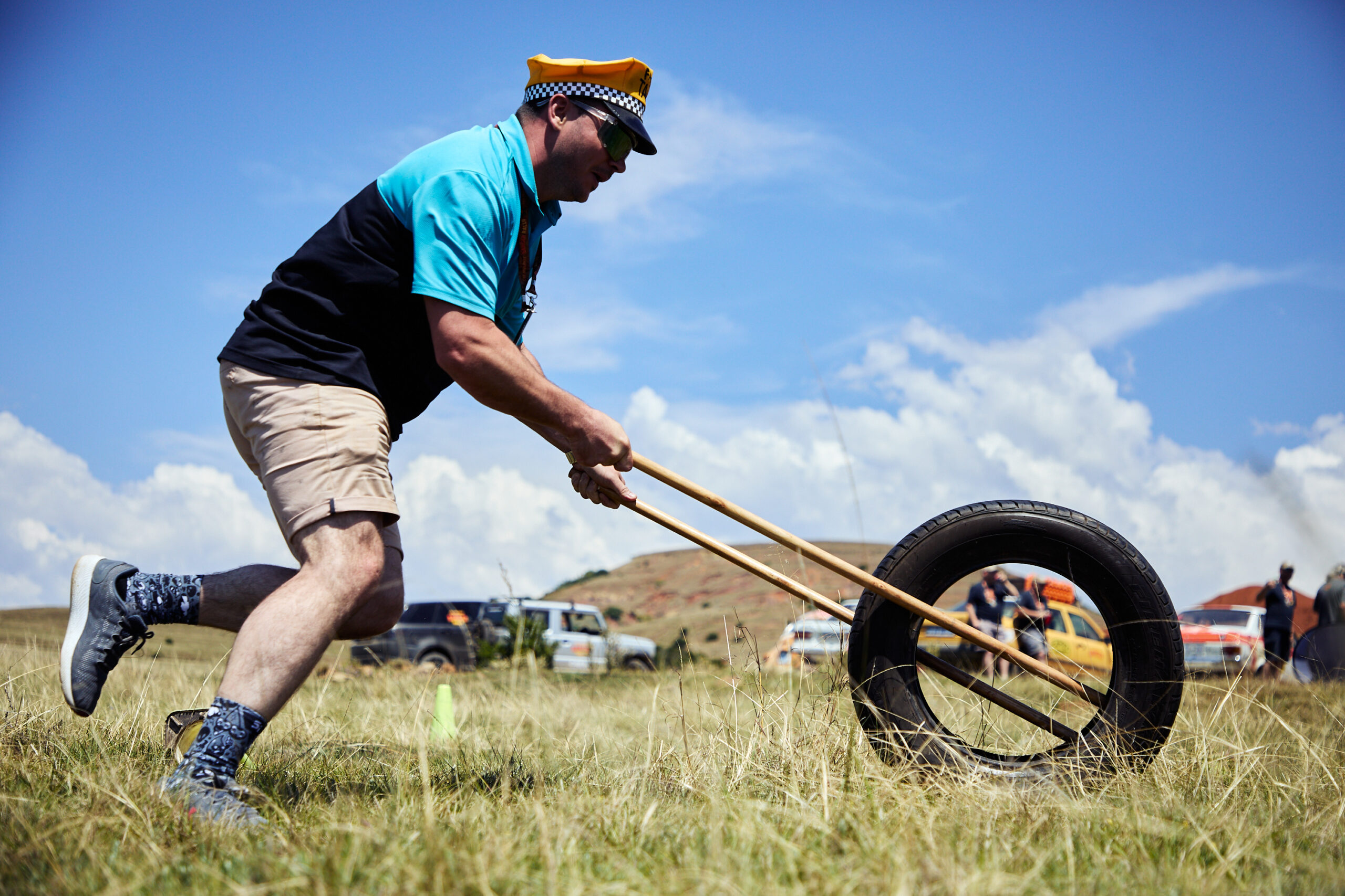 A participant in a yellow taxi hat competes in a tire-rolling out-of-car challenge during the CBR Midlands Banger Rally in a grassy field.